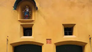 Eine gelbe Hausfassade mit einer Heiligenfigur in einer Nische über zwei grünen Türen.
