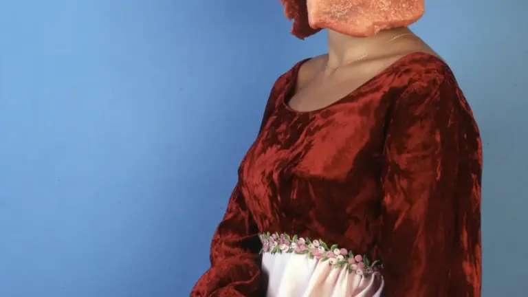 Eine Frau in einem historischen Kleid trägt rohes Fleisch als Kopfbedeckung vor blauem Hintergrund.
