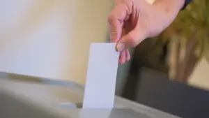 Eine Hand wirft einen leeren weißen Wahlzettel in eine graue Wahlurne aus Kunststoff.