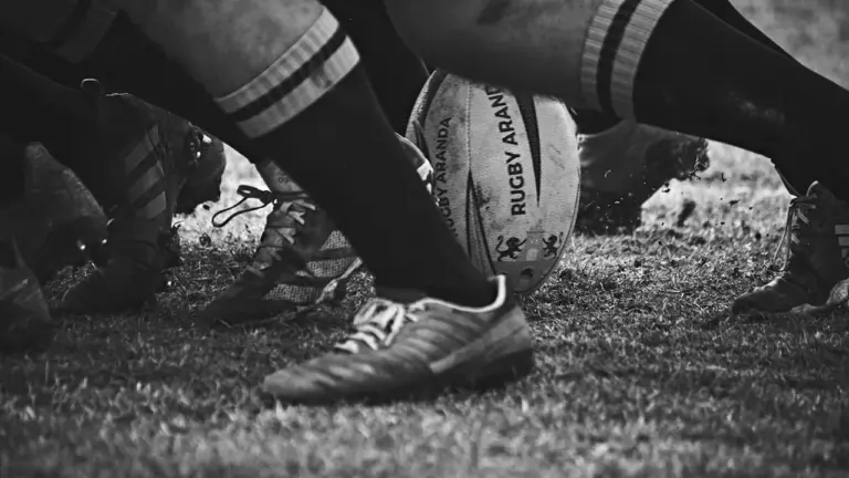 Schwarz-Weiß-Aufnahme eines Rugbyballs im Zentrum eines intensiven Spielgeschehens auf dem Rasen.