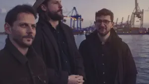 Drei Männer stehen vor einer Hamburger Hafen-Kulisse mit Kränen und Wasser bei einsetzender Abenddämmerung.
