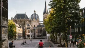 Blick auf den Aachener Dom mit umliegendem gepflastertem Platz und entspannten Passanten bei Sonnenuntergang.