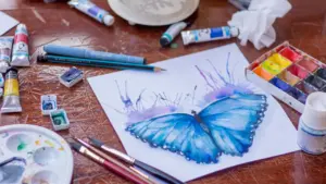 Ein Aquarellbild eines blauen Schmetterlings umgeben von verschiedenen Malutensilien auf einem Holztisch.