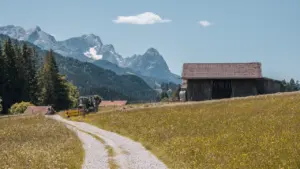 Ein idyllischer Feldweg führt zu einer rustikalen Scheune vor einer malerischen Bergkulisse.