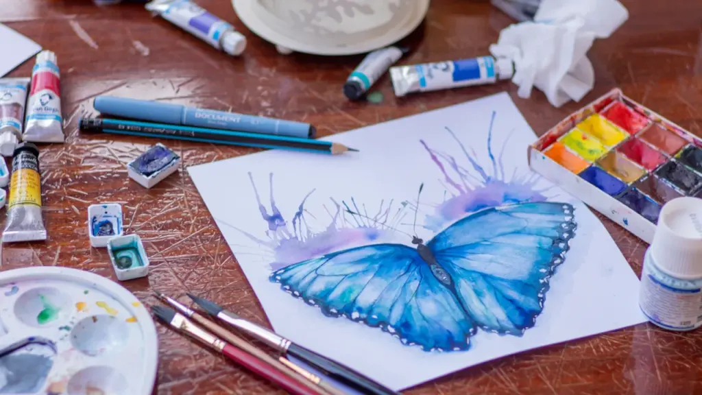 Ein Aquarellbild eines blauen Schmetterlings umgeben von verschiedenen Malutensilien und Farben.