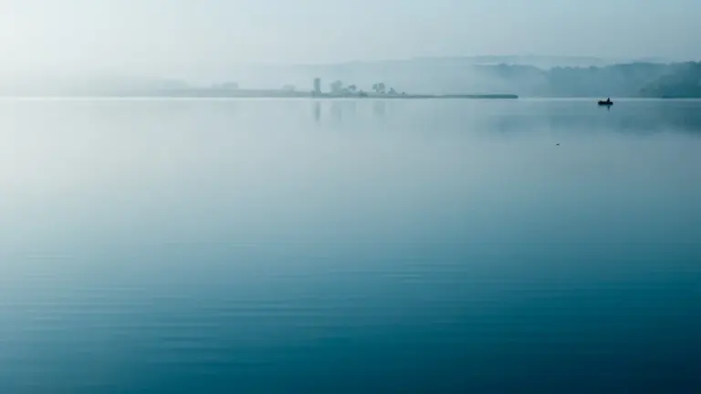 Ein ruhiger, nebliger See am frühen Morgen in einer friedlichen, atmosphärischen Landschaft.